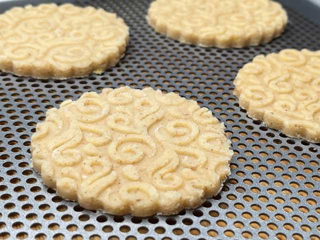 Biscotti crudi di pasta frolla alla chufa con decorazioni a spirale, disposti su una teglia microforata prima della cottura.