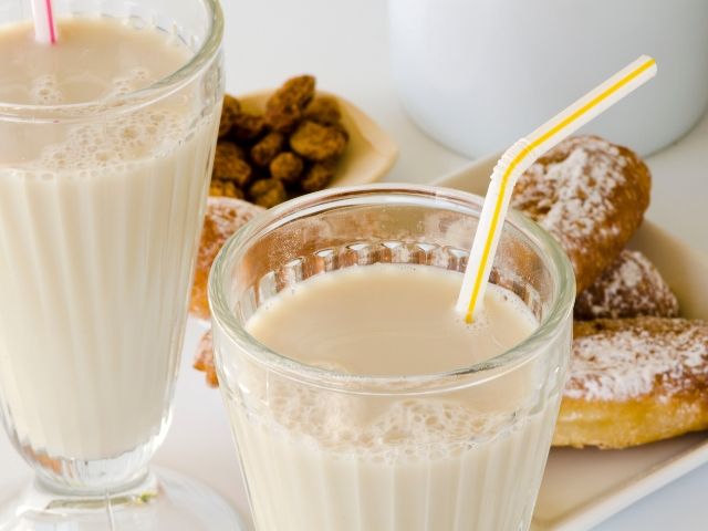 Bicchieri di bevanda color crema a base di chufa con dolcetti sullo sfondo.